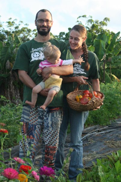 A young couple with a todder, holding a basket of heirloom tomatoes, stands in a field of banana trees and colorful zinnia flowers.