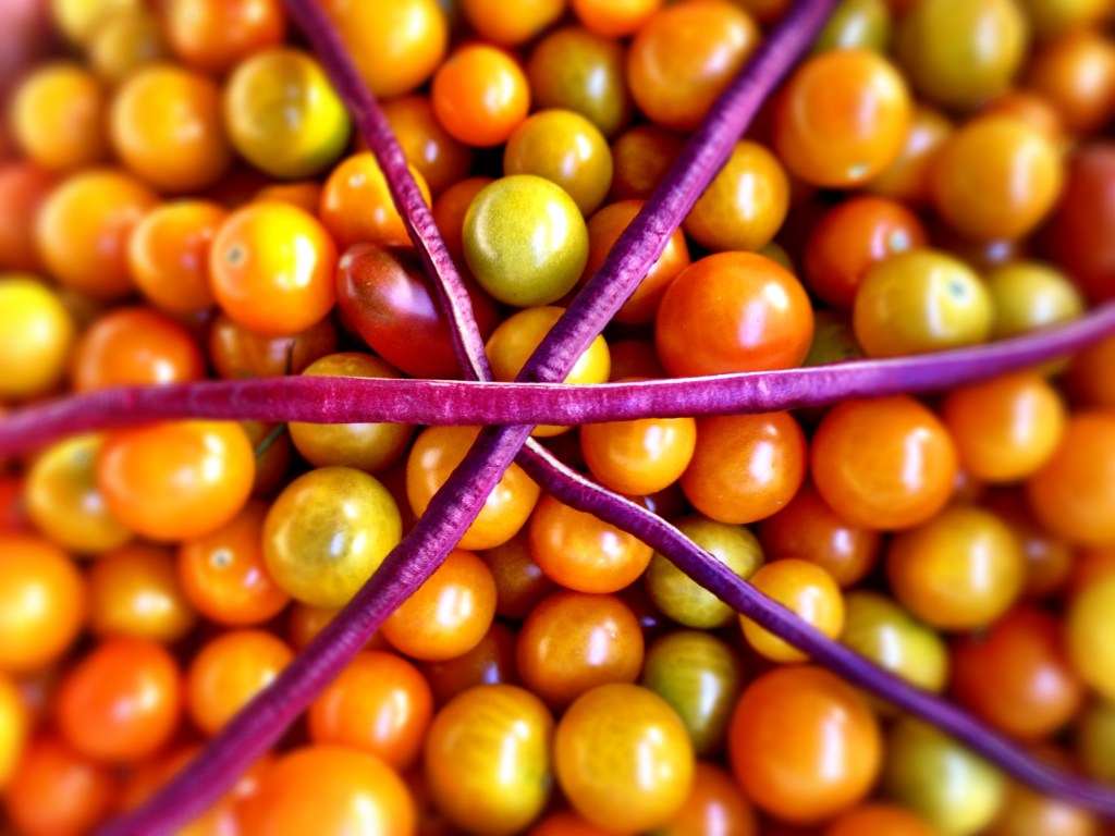 Three purple longbeans intersect to form a star on top of a pile of ripe yellowy-orange cherry tomatoes.